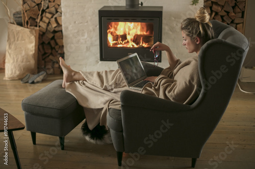 Beautiful middle-aged woman next to the fireplace relaxes in the living room and works on laptop from house. Girl in 30's surveyed for the  home bussiness project