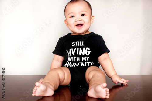 A baby boy is wearing a shirt with a message saying he is still living with my parents