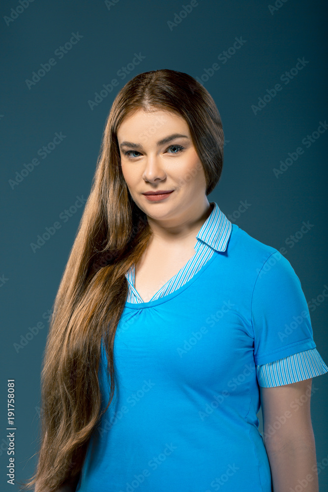 Attractive plus-sized woman wearing blue blouse in studio setting. Girl with long hair over blue background Attractive plus-sized woman wearing blue blouse in studio setting. Girl with long hair over blue background