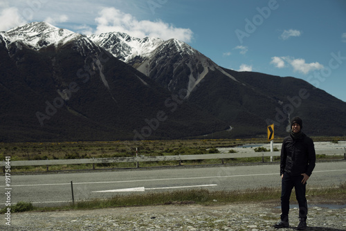 Hombre joven posando frente a paisaje con carretera y montañas nevadas con cielo azul nuboso.