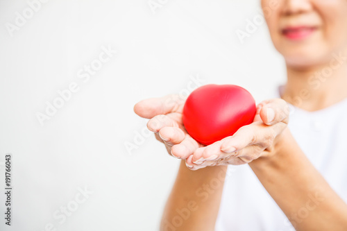 Fototapeta Naklejka Na Ścianę i Meble -  Red heart held by smiling female nurse's hand, representing all effort to deliver high quality service mind to patient. Professional, Specialist in white uniform isolated on white background.