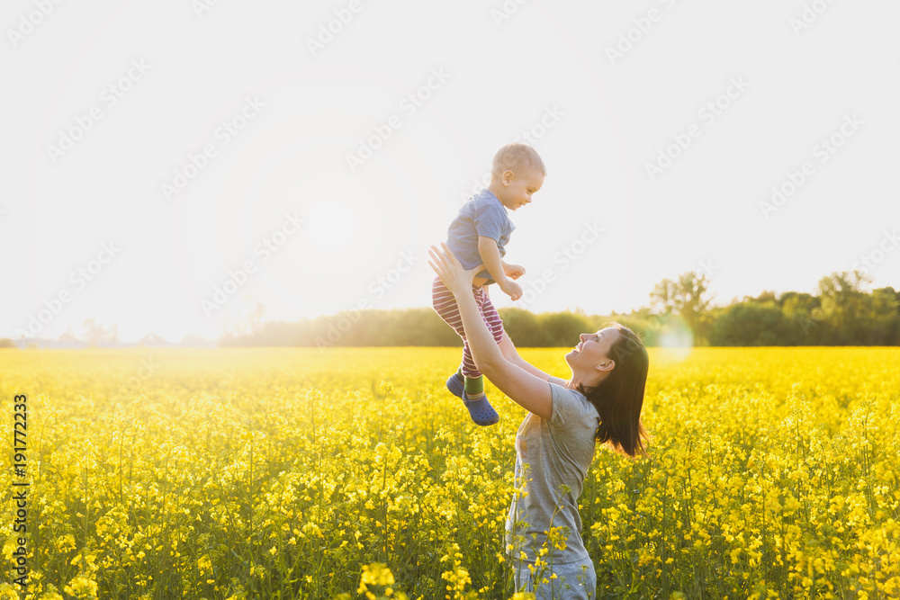 Fototapeta premium Joyful woman walk on green yellow flowering field background, rest, have fun, play, toss up little cute child baby boy. Mother, little kid son. Family day 15 of may, love, parents, children concept.