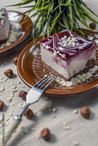 A piece of vanilla cake with chocolate base and cherry glaze decorated with some pieces of white chocolate served in a brown clay dish with silver fork placed on a beige tablecloth. Blurred background
