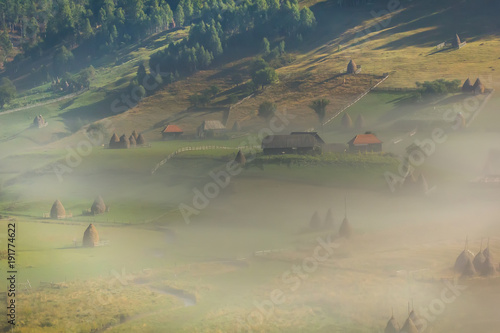 Beautiful rural mountain landscape in the morning light with fog, old houses and haystacks, Fundatura Ponorului, Hunedoara County, Romania photo