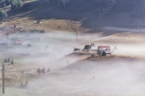 Beautiful rural mountain landscape in the morning light with fog, old houses and haystacks, Fundatura Ponorului, Hunedoara County, Romania