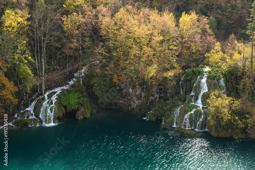 An aerial picture of two waterfall and a hiking trail in Plitvice Lakes National Park. The trees are losing leaves and changing colors in the fall. The water is a turquoise blue and the grass is lush.