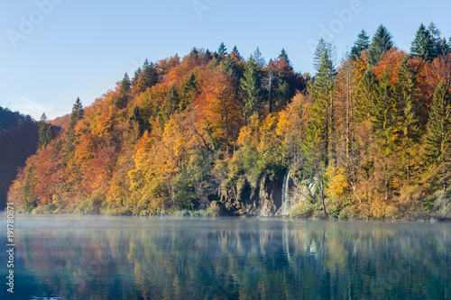 Fototapeta Naklejka Na Ścianę i Meble -  A waterfall flows into a turquoise lake in autumn. The trees are red orange green yellow and the sky is clear. There is a layer of mist or fog on top of the still water.