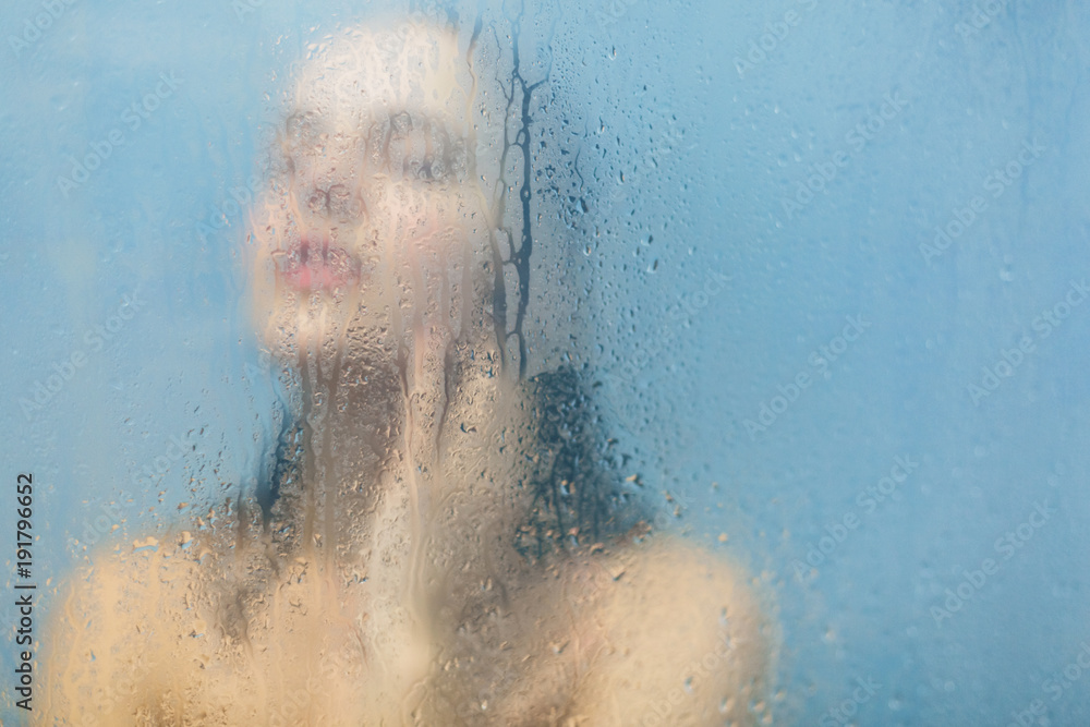 Unfocused silhouette of naked female model takes shower in cabine feels relaxed and takes care of her body, woman washes behind weeping glass shower door. Taking bath. Skin care concept