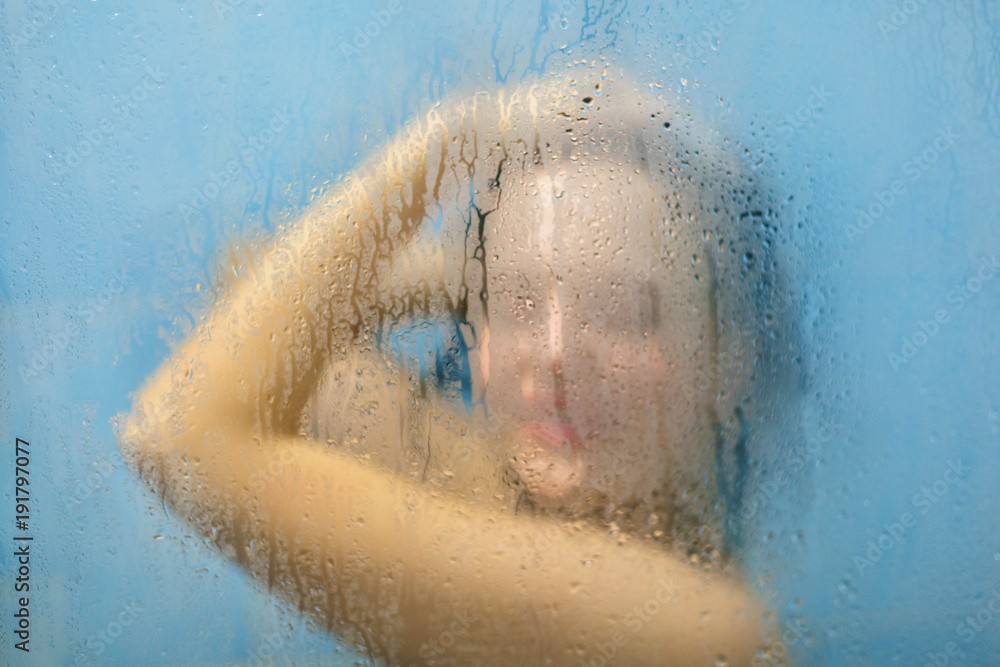 Fototapeta premium Young woman refreshes in shower, pamper her skin with hot water, poses in shower cabine with blurred sweat glass. Attractive young female feels relaxation as bathes in bathroom alone