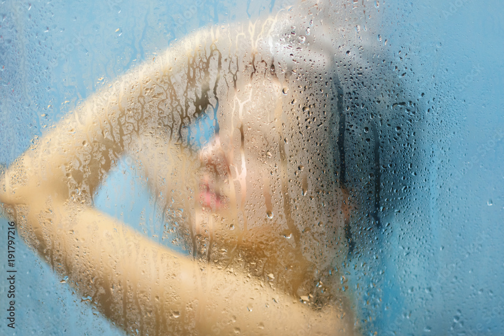 Fototapeta premium Indoor shot of naked female takes shower at bathroom, washes hair, feels relief after hard working day, likes cleannless, takes care of body and skin. Unfocused sillouhette of woman in shower cabine