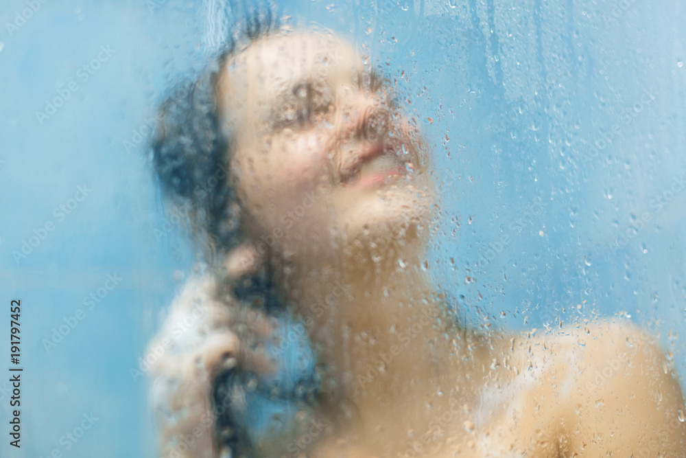 Single pleased young woman takes shower in bathroom, washes hair, takes care of her beauty and appearance, has positive smile. Blurred background with water drops and sweat. Relax concept