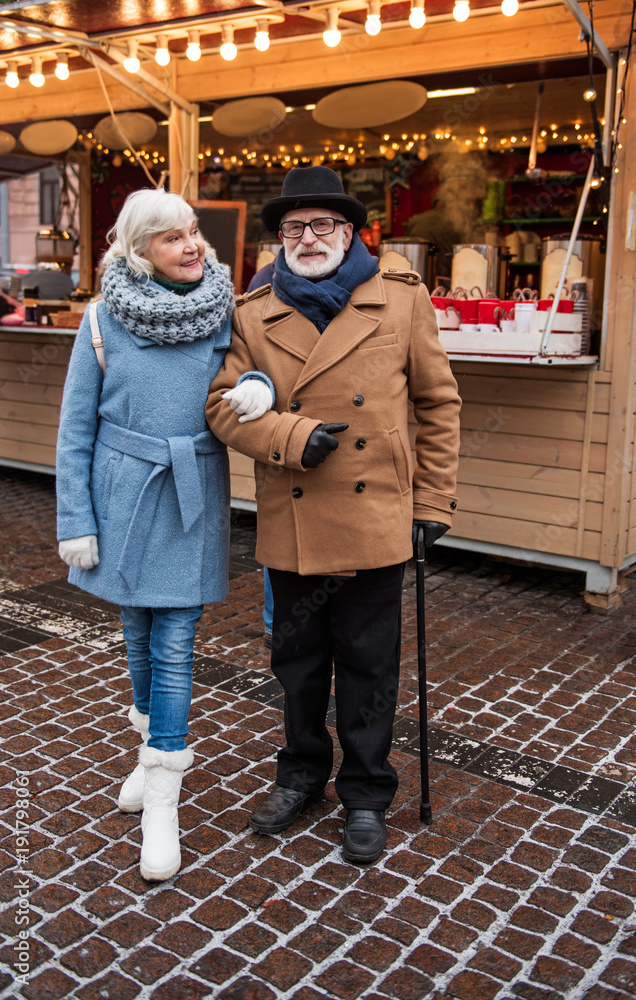 Obraz premium Full length portrait of cheerful senior couple standing on street arm in arm. Confectionary shop is on background