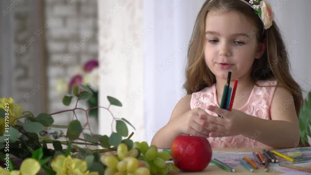 A girl in a pink dress is laying out colorful pencils on a table with an apple and grapes A girl in a pink dress is laying out colorful pencils on a table with an apple and grapes