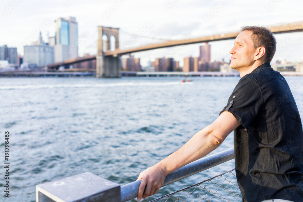 Back of young man outside outdoors in NYC New York City Brooklyn Bridge Park by east river, railing, looking at view of cityscape skyline