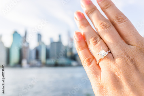 Young woman's hand with diamond engagement ring princess cut, gold outside outdoors in NYC New York City Brooklyn Bridge Park by east river, cityscape, skyline bokeh