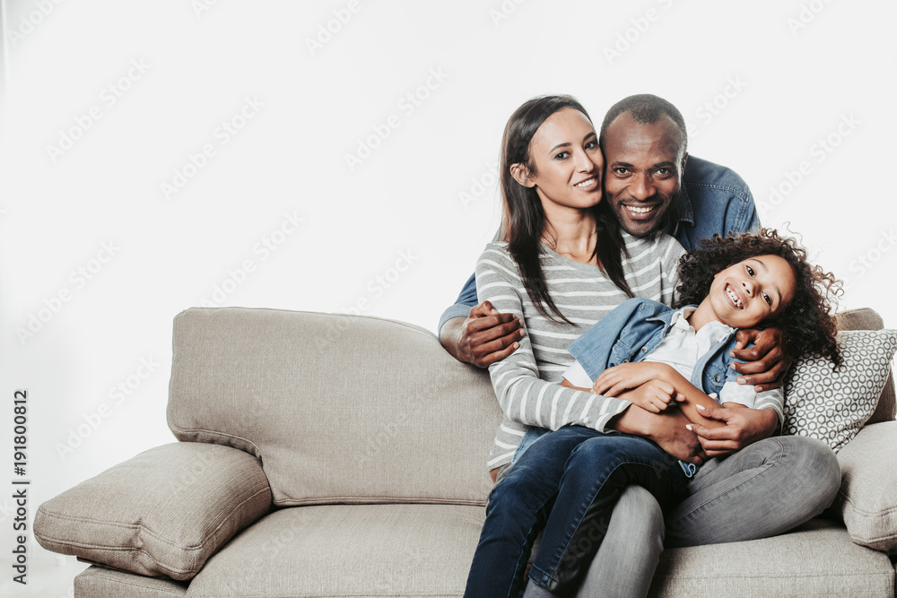 Portrait of glad parents and daughter spending time together. Kid is sitting at moms feet and father embracing them aback. Copy space in left side. Isolated on background Portrait of glad parents and daughter spending time together. Kid is sitting at moms feet and father embracing them aback. Copy space in left side. Isolated on background