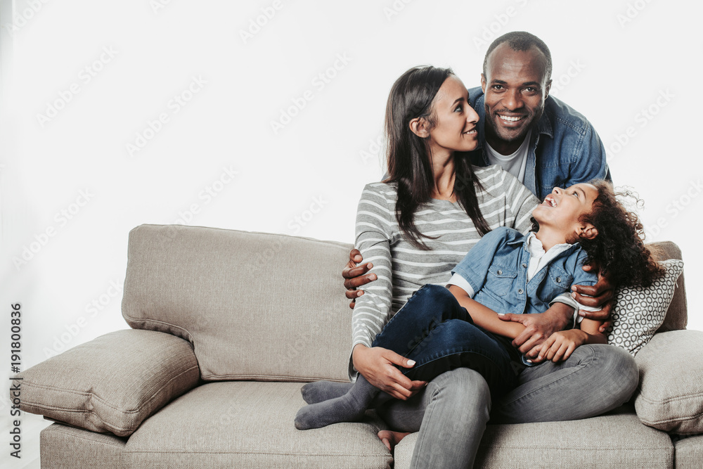 Enjoyed parents and child sitting together with comfort. Mom and kid are looking at father standing abaft them. Copy space in left side. Isolated on background Enjoyed parents and child sitting together with comfort. Mom and kid are looking at father standing abaft them. Copy space in left side. Isolated on background
