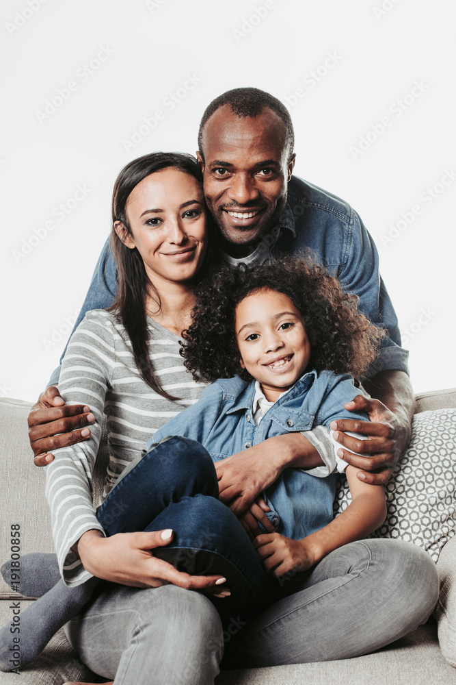 Portrait of happy mother, father and daughter resting on divan and looking at camera. Girl is on mums knees and dad is hugging them. Isolated on background Portrait of happy mother, father and daughter resting on divan and looking at camera. Girl is on mums knees and dad is hugging them. Isolated on background