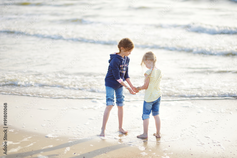 Two little sisters having fun on a sandy beach on warm and sunny summer day. Kids playing by the ocean. Two little sisters having fun on a sandy beach on warm and sunny summer day. Kids playing by the ocean.