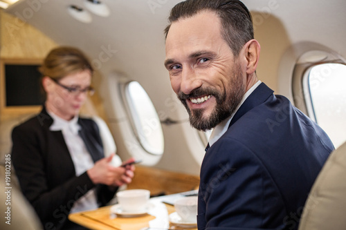 Portrait of contented middle aged male in airplane with his colleague sitting in front of him, he is looking at camera with broad smile. Focus on man