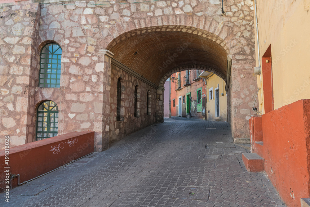 Fototapeta premium A stone paved street going up a hill and through a stone tunnel with two arched windows on the stone wall, next to the tunnel entrance, and colorful houses, in Guanajuato, Mexico