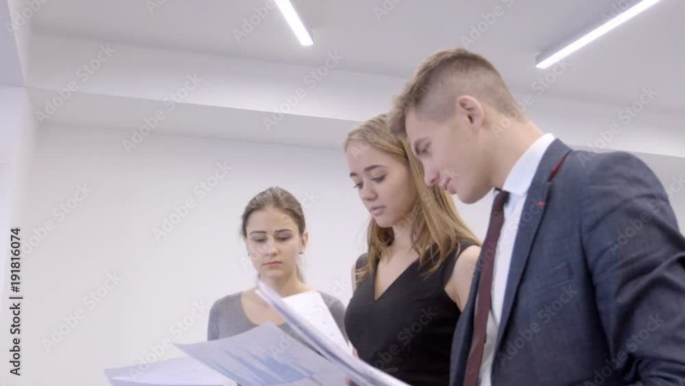Three traiders are researching paper reports with digital currency market situation in modern office. Businessman and two female financial analytics are standing in a row and looking to the documents Three traiders are researching paper reports with digital currency market situation in modern office. Businessman and two female financial analytics are standing in a row and looking to the documents
