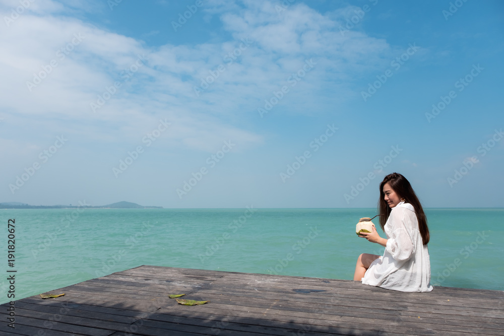 A beautiful asian woman on white dress sitting at the terrace drinking coconut juice with sea and blue sky background with feeling relaxed