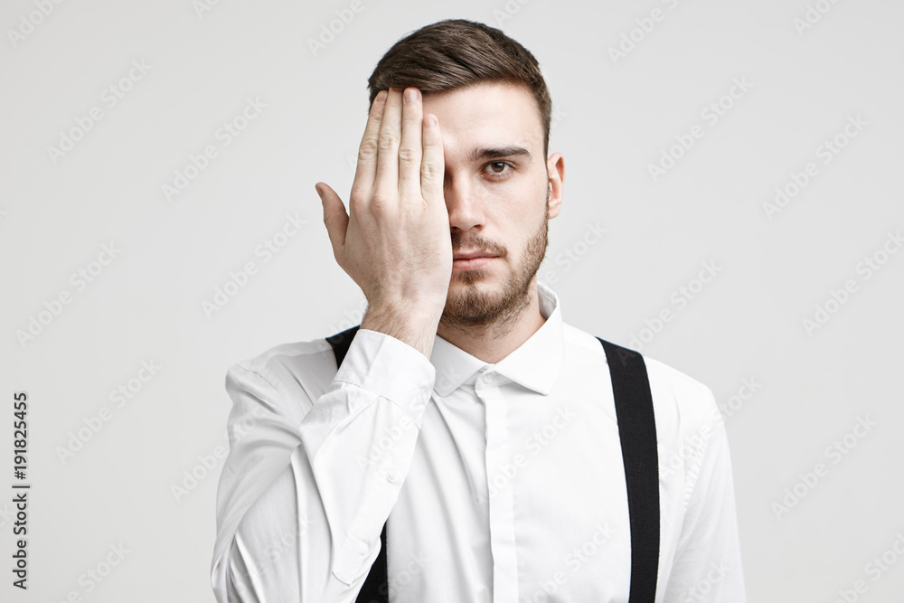 Isolated studio shot of handsome young male corporate worker with bristle and stylish haircut looking at camera, covering one eye with palm as if having his eyes tested during vision examination Isolated studio shot of handsome young male corporate worker with bristle and stylish haircut looking at camera, covering one eye with palm as if having his eyes tested during vision examination