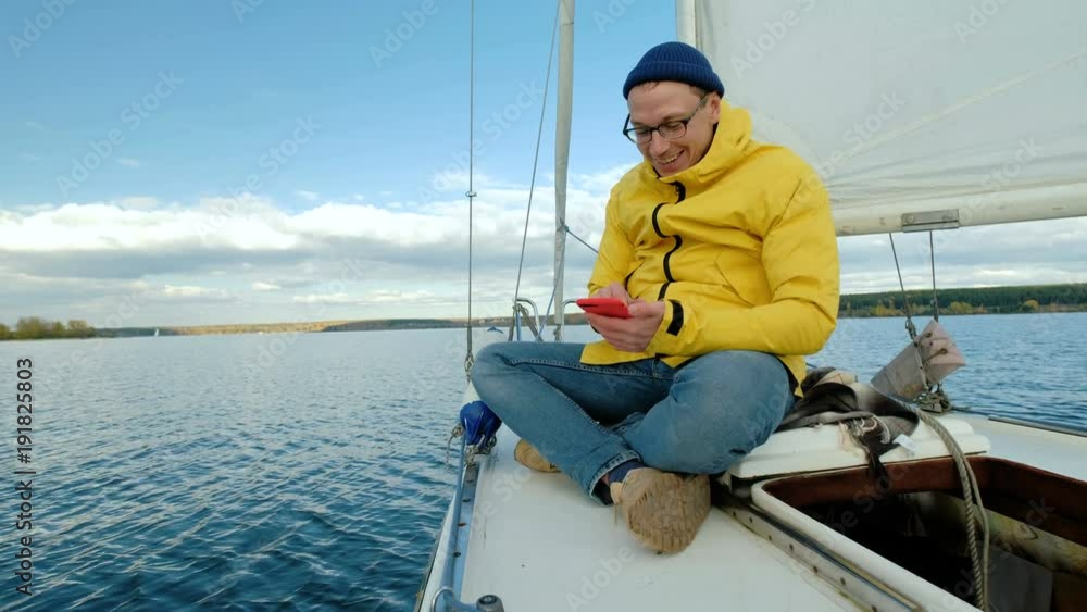 Young man sitting on sailboat and browsing smartphone on background of sea Young man sitting on sailboat and browsing smartphone on background of sea