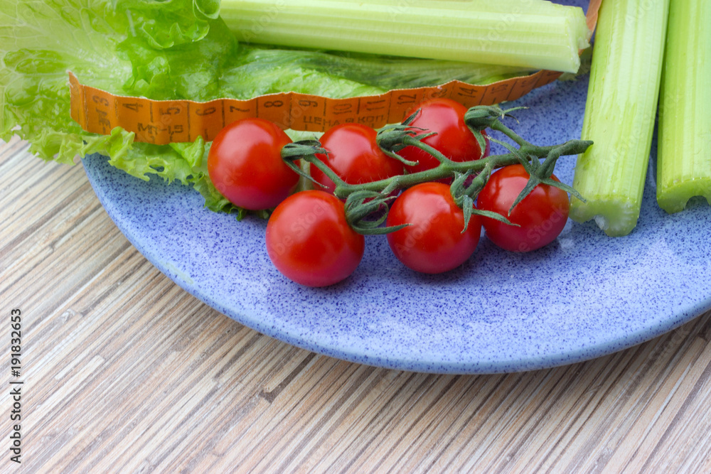 big celery,tape measure for diet, fresh green lettuce leaves next to red pepper and red cherry tomatoes lying on a blue plate, wooden table background big celery,tape measure for diet, fresh green lettuce leaves next to red pepper and red cherry tomatoes lying on a blue plate, wooden table background