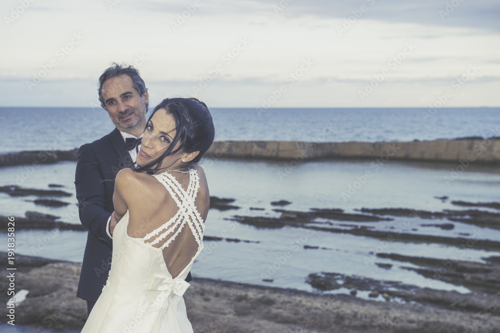 A newly married couple is embracing . On the background, sea and rocks of the wonderful location of Plemmirio natural park in Siracusa (Sicily), .