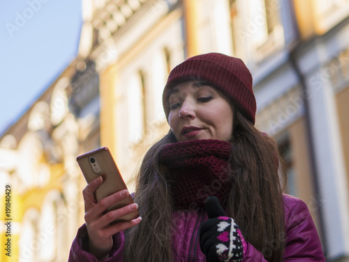 Pretty Brunette Girl Wearing Purple Winter Coat, Hat and Scarf, Walking by European Street at Winter, make Photos on Her Smartphone and Making Selfie photo