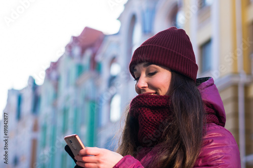 Pretty Brunette Girl Wearing Purple Winter Coat, Hat and Scarf, Walking by European Street at Winter, make Photos on Her Smartphone and Making Selfie photo