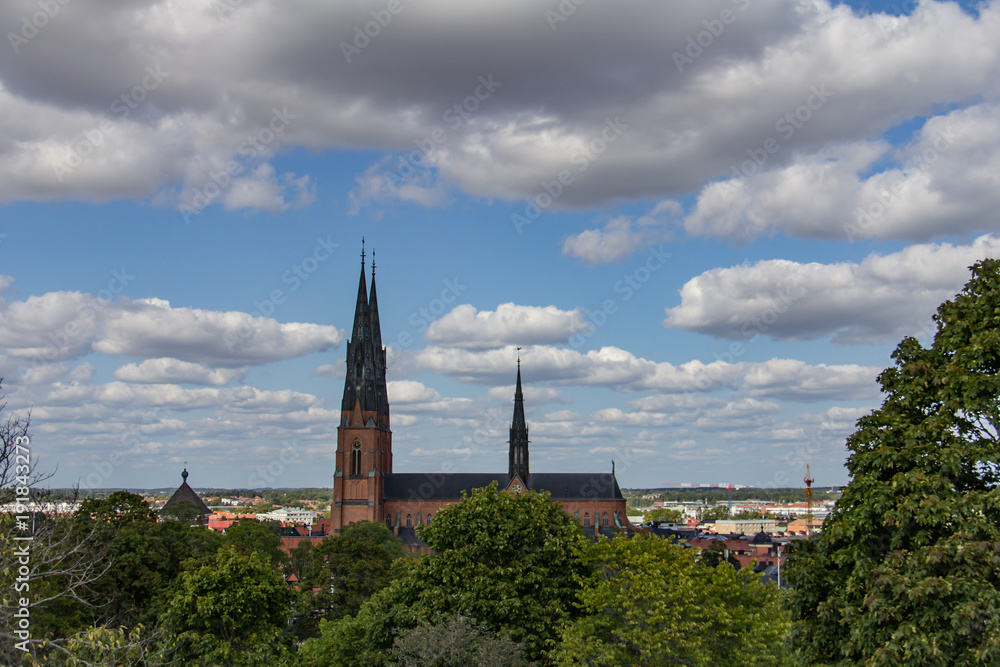 Profile of Uppsala Cathedral in Sweden under a blue sky with fluffy white clouds, green trees in the foreground Profile of Uppsala Cathedral in Sweden under a blue sky with fluffy white clouds, green trees in the foreground