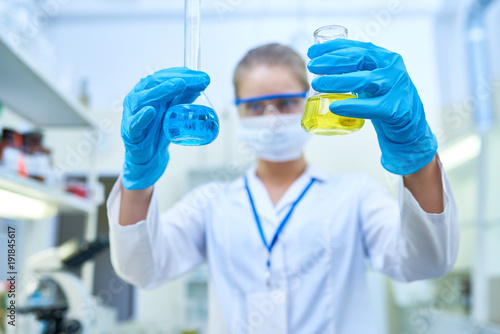 Portrait of female scientist wearing blue protective gloves holding test tubes with colored liquids while studying chemicals in medical laboratory, focus on hands