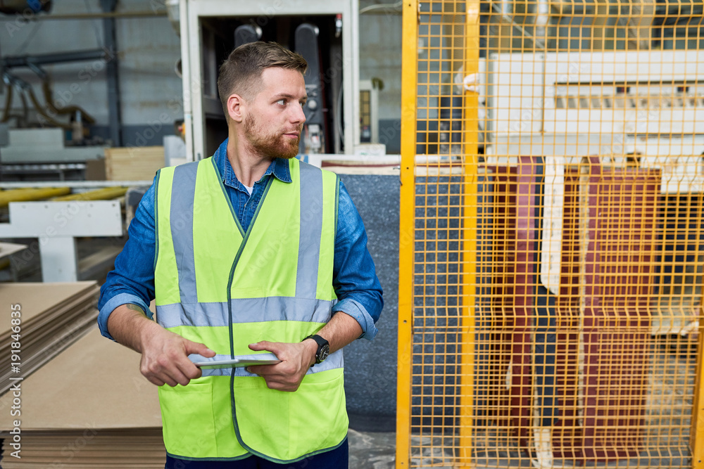 Portrait of young bearded factory worker wearing reflective jacket posing in industrial workshop looking away and holding digital tablet, copy space Portrait of young bearded factory worker wearing reflective jacket posing in industrial workshop looking away and holding digital tablet, copy space