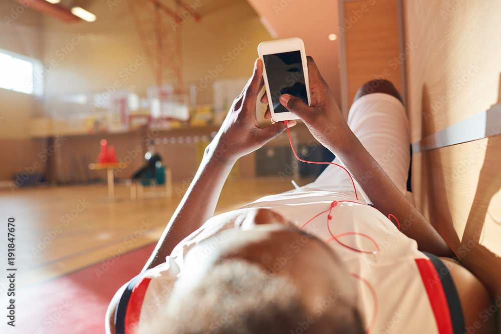 Naklejka premium Tired African basketball player waiting for training and relaxing on bench enjoying music in earphones and using gadget while checking social media on training court