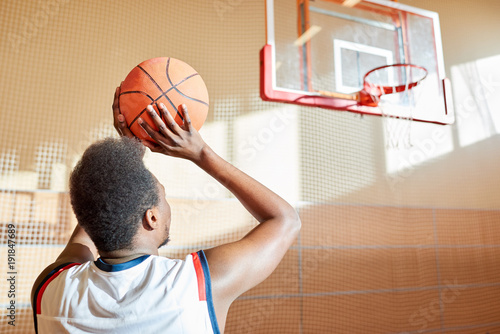 Rear view of ambitious African-American sportsman preparing for score aiming before throwing ball in basket on basketball court
