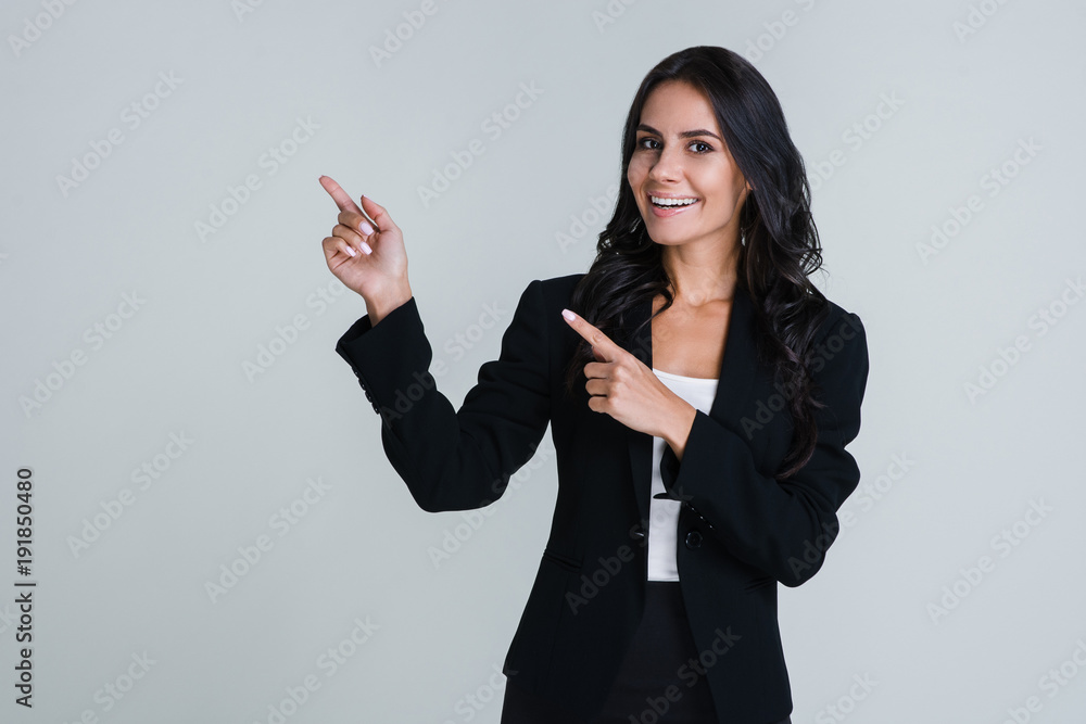 Look here! Beautiful young businesswoman pointing away and looking at camera with smile while standing against white background Look here! Beautiful young businesswoman pointing away and looking at camera with smile while standing against white background