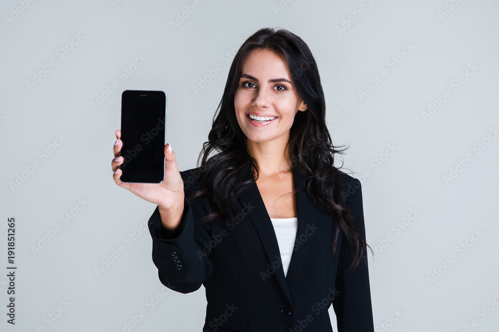 Have you seen this app? Beautiful young businesswoman showing her smartphone and looking at camera with smile while standing against white background - obrazy, fototapety, plakaty 