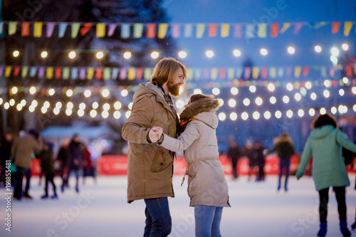 Ice skating rink and lovers together. A pair of young people in an embrace on a city skating rink lit by light bulbs and bright lights. Winter date for Christmas on the ice arena