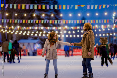 Theme ice skating rink and loving couple. meeting young, stylish people ride by hand in crowd on city skating rink lit by light bulbs and lights. Ice skating in winter for Christmas on ice arena