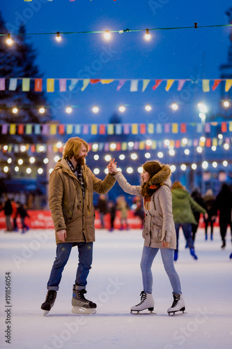 Theme ice skating rink and loving couple. meeting young, stylish people ride by hand in crowd on city skating rink lit by light bulbs and lights. Ice skating in winter for Christmas on ice arena