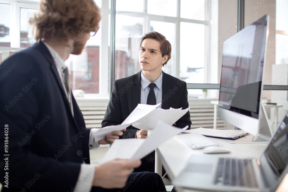 Two young bankers sitting in office and discussing papers and financial information Two young bankers sitting in office and discussing papers and financial information