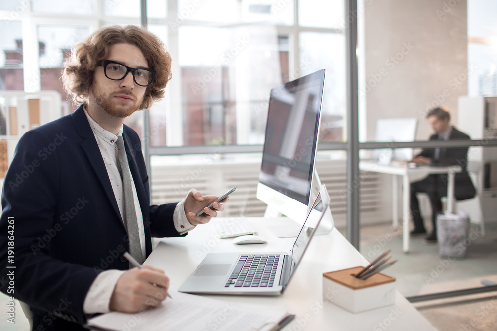 Young serious office worker with smartphone and laptop messaging by his workplace Young serious office worker with smartphone and laptop messaging by his workplace
