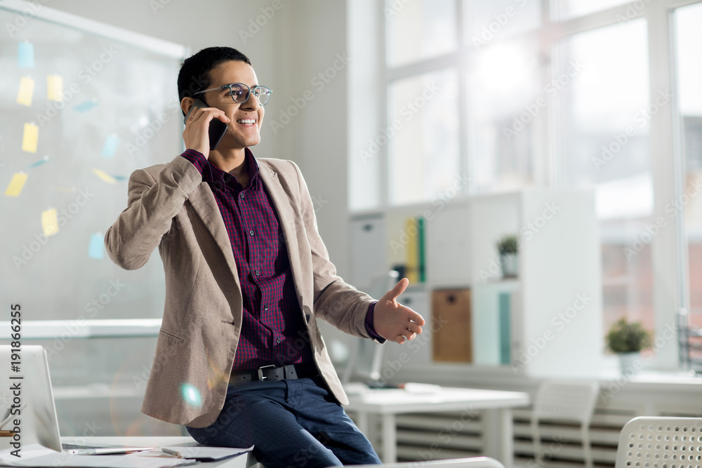 Successful broker in elegant formalwear sitting on desk and consulting one of his clients by the phone Successful broker in elegant formalwear sitting on desk and consulting one of his clients by the phone