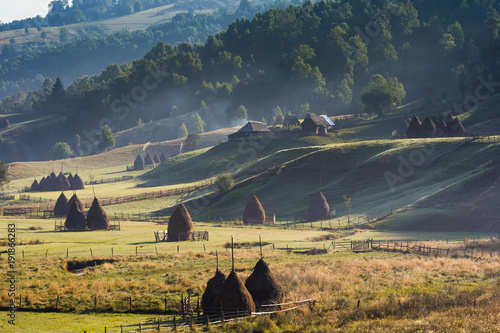 Beautiful rural mountain landscape in the morning light with fog, old houses and haystacks, Fundatura Ponorului, Hunedoara County, Romania photo