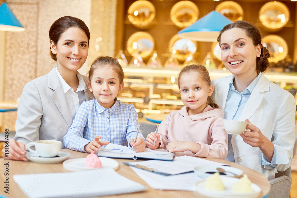 Two young businesswomen and their daughters sitting in cafe and having drinks and rest