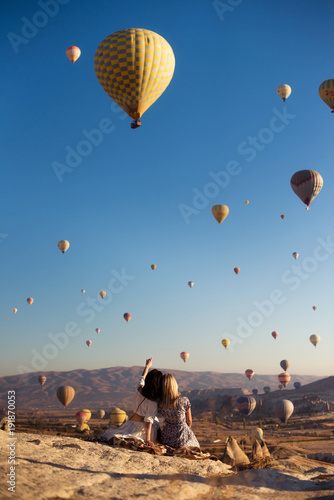 Принти на полотні Outdoor view of two female best friends embrace each other, have good relationship, admire beautiful landscapes, sit together on high hill, look at parachutes and blue sky