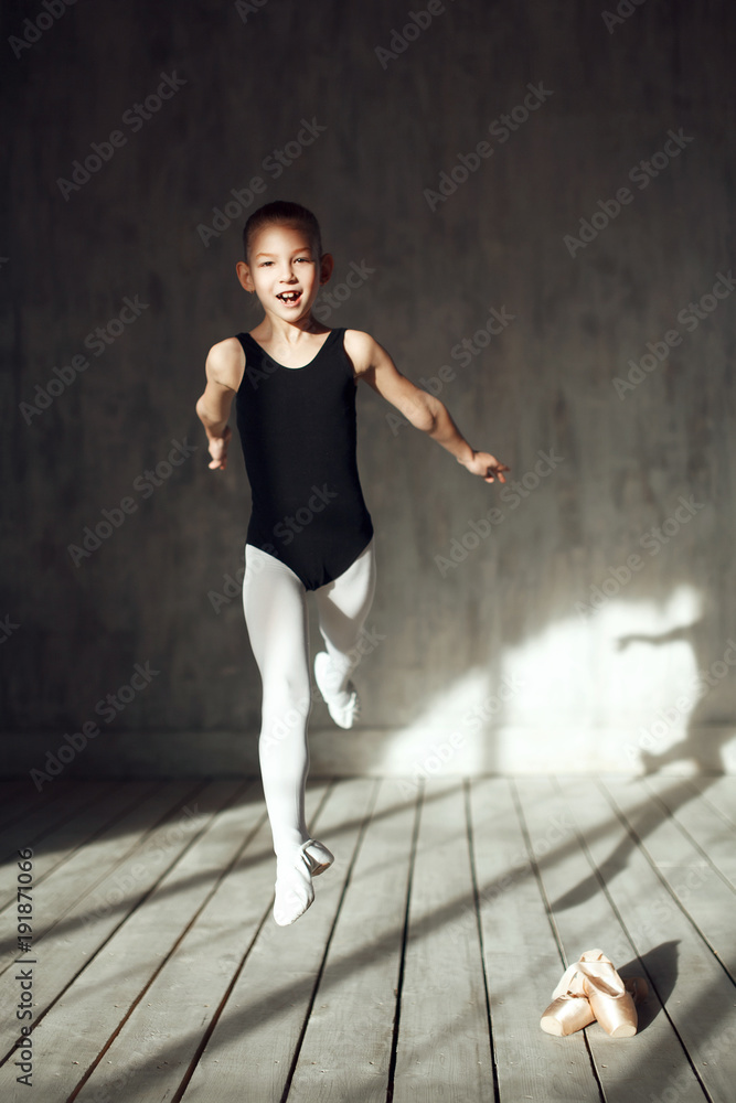 A little adorable young ballerina in black sportwear and white tights jumps  in the interior studio posing on camera in bright sunlight from window  Stock Photo